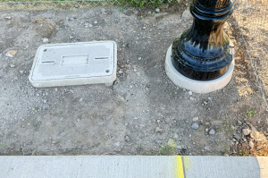 A close-up of a utility box lid set into the ground next to the base of a decorative streetlight pole in Herriman, Utah. The utility box is surrounded by dirt and gravel, with part of a newly finished sidewalk visible at the bottom of the frame. The base of the streetlight is labeled "Herriman" in raised text.